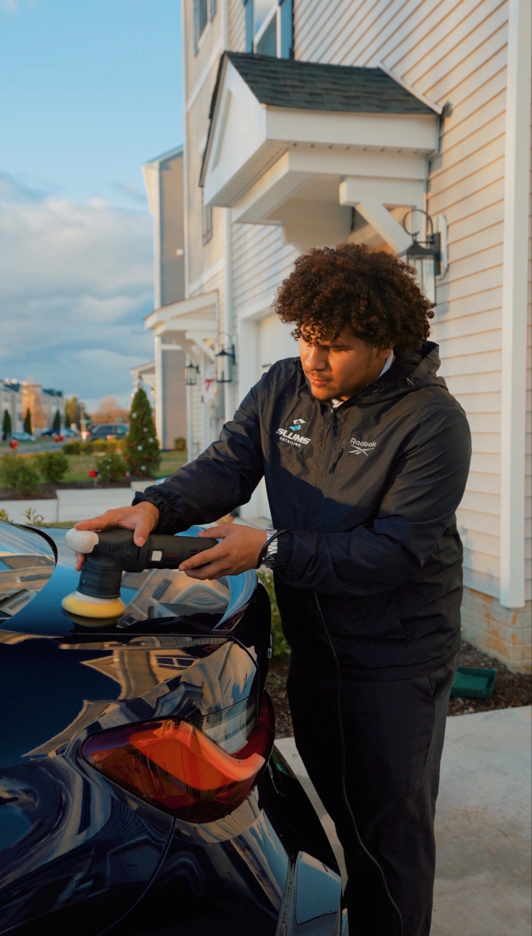 Owner-operator polishing a black car at sunset with Slums Mobile Detailing branding